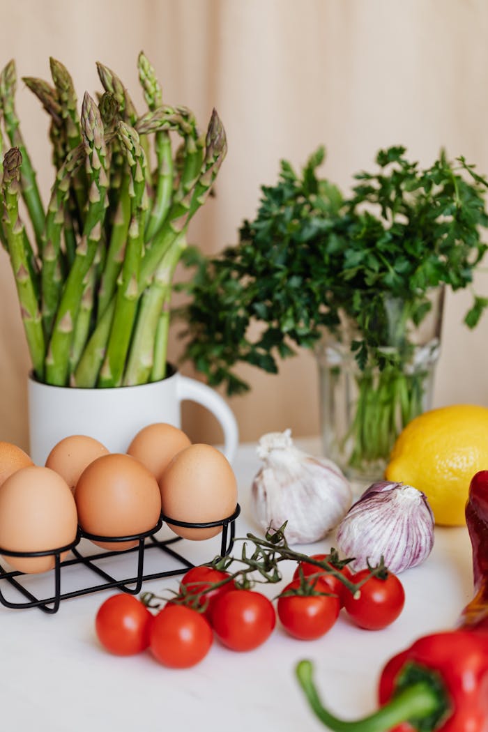 Assorted fresh vegetables with eggs and lemon arranged on table near bunch of parsley in glass and asparagus in white cup on kitchen